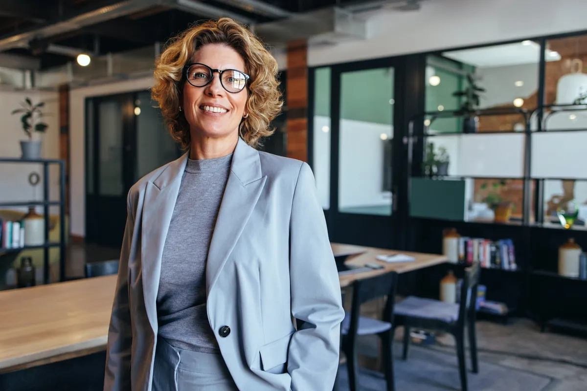 A smiling businesswoman in glasses and a gray suit stands in a modern office