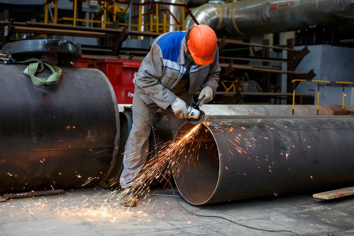 A pipefitter in an orange hard hat grinds the edge of a large steel pipe, sending sparks across the shop floor